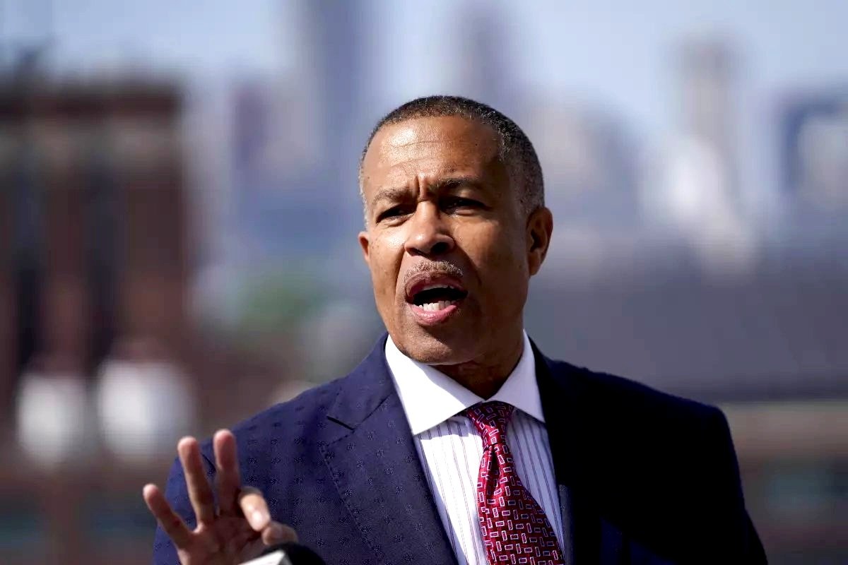 James Craig in suit and red tie speaking outdoors, blurred cityscape background.